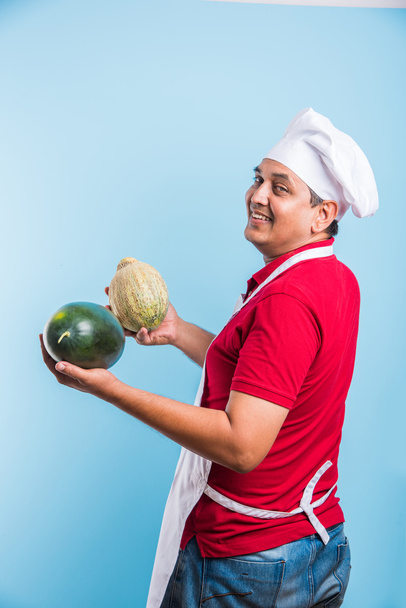 handsome indian male chef holding water melon and musk melon, asian male chef holding fruits, isolated over blue background - Photo, Image
