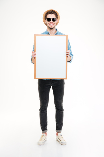 Smiling young man in hat and sunglasses holding blank whiteboard - Photo, Image