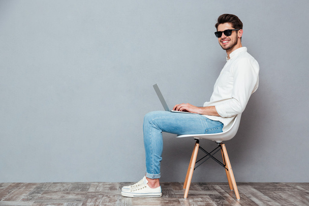 Smiling young man in white shirt and sunglasses using laptop - Фото, зображення