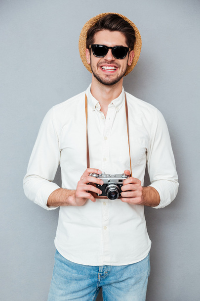 Cheerful young man in hat and sunglasses holding old camera - Foto, Imagen