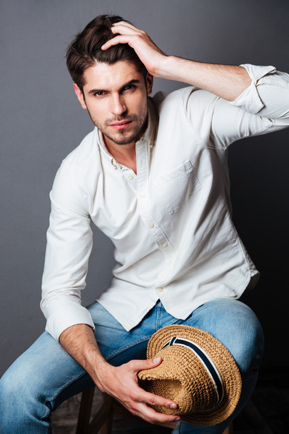 Fashion portrait of attractive young man sitting and holding hat - Foto, Imagem