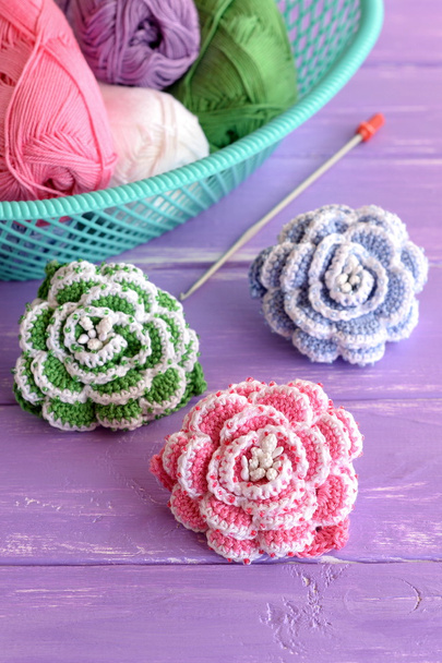 Ροζ, πράσινο και γαλάζιο πλέκω λουλούδια που διακοσμούνται με χάντρες. Βαμβακερά νήματα κούκλες στο καλάθι, γάντζο και φωτεινά πλεκτό τριαντάφυλλα σε λιλά φόντο ξύλινη - Φωτογραφία, εικόνα