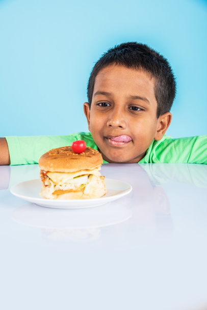 χαριτωμένο Ινδικό αγόρι και burger σε μικρό λευκό πλάκα, μικρό ασιατικό αγόρι και burger, πάνω από πολύχρωμο φόντο - Φωτογραφία, εικόνα