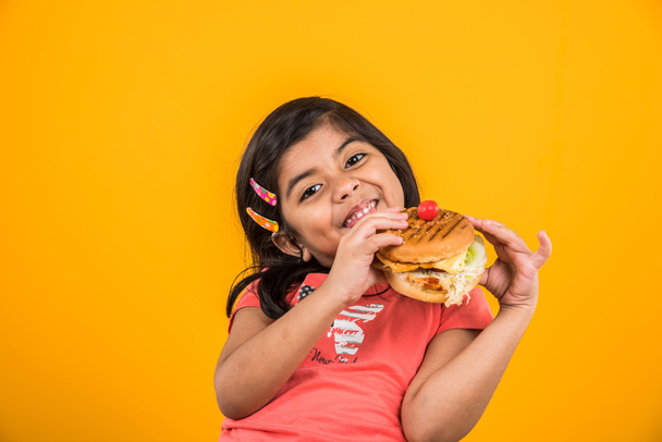 linda chica india comiendo hamburguesa, pequeña chica asiática y hamburguesa, aislado sobre fondo amarillo
 - Foto, Imagen