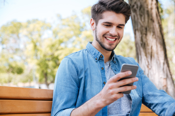 Smiling young man using smartphone sitting on the bench outdoors - Foto, immagini
