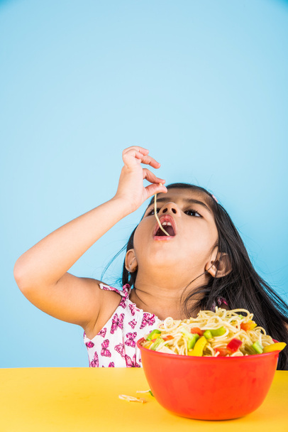 Счастливый азиатский ребенок ест вкусную лапшу, маленькая индийская девочка ест лапшу в красной миске
 - Фото, изображение