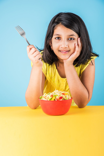 Счастливый азиатский ребенок ест вкусную лапшу, маленькая индийская девочка ест лапшу в красной миске, на голубом фоне
 - Фото, изображение