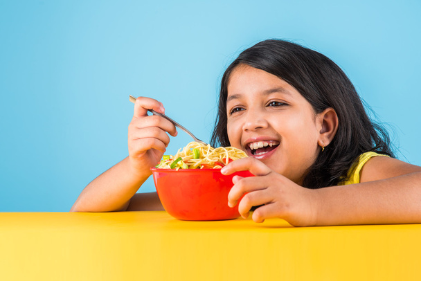 Happy Asian child eating delicious noodle, small indian girl eating noodles in red bowl, over blue background - Photo, Image