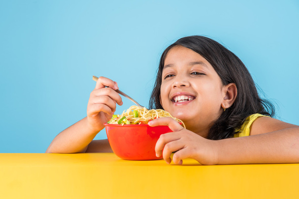 Niño asiático feliz comiendo fideos deliciosos, niña india pequeña comiendo fideos en tazón rojo, sobre fondo azul
 - Foto, Imagen