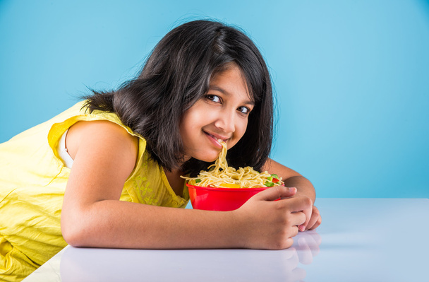 glückliches asiatisches Kind isst leckere Nudeln, kleines indisches Mädchen isst Nudeln in roter Schüssel, vor blauem Hintergrund - Foto, Bild