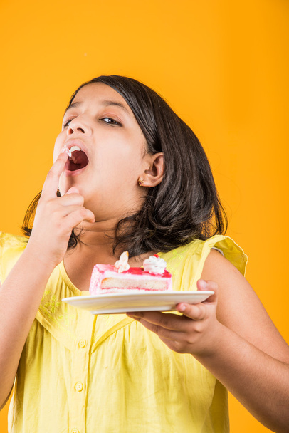 portrait of Indian kid eating cake or pastry, cute little girl eating cake, girl eating strawberry cake over yellow background - Photo, Image
