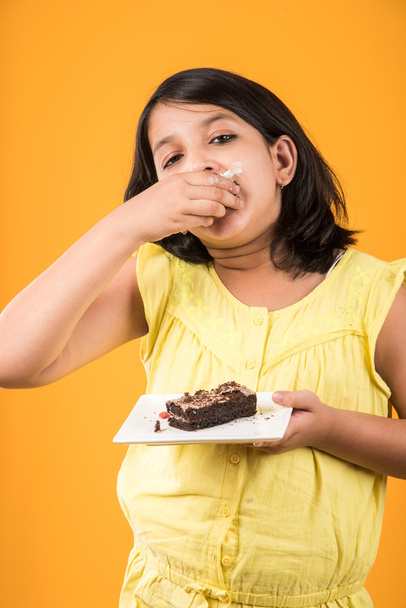 Porträt eines indischen Kindes, das Kuchen oder Gebäck isst, nettes kleines Mädchen, das Kuchen isst, Mädchen, das Schokoladenkuchen oder Gebäck vor buntem Hintergrund isst - Foto, Bild