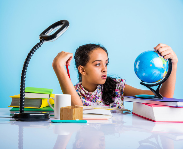 indian girl student looking at educational globe and sitting at table with books, table lamp and milk mug, asian girl child studying geography, curious asian girl studying geography with globe - Photo, Image