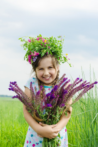 Piccola sorridente ragazza felice con un mazzo di fiori selvatici e una ghirlanda in testa tra i prati verdi
  - Foto, immagini