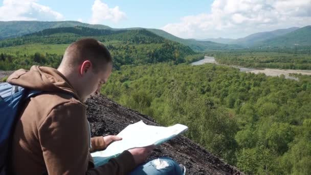 Young active man checking landscape terrain with map and compass while sitting on rocky mound - Felvétel, videó