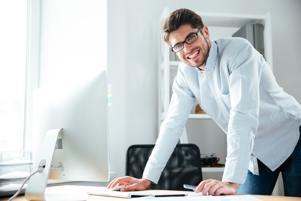 Cheerful young businessman standing and using computer in office - Foto, Imagem