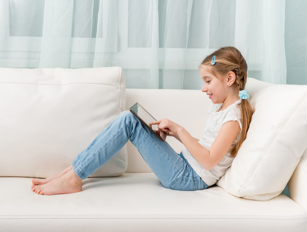 little girl sitting on white sofa looks at tablet touching it - Фото, изображение
