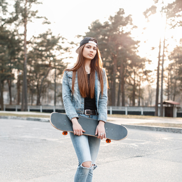 Beautiful woman in cap and jeans clothes holding a skateboard in a spring sunny day. - Photo, Image