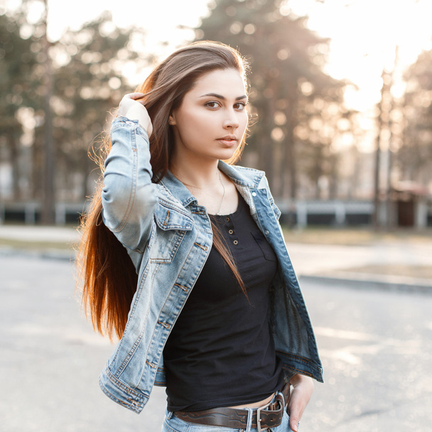 Stylish young girl in fashionable jeans clothes corrects her long hair at sunset - Photo, image