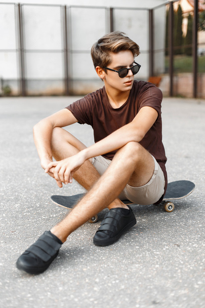Handsome young man with the hairstyle in fashion sunglasses sitting on a skateboard. - Photo, Image