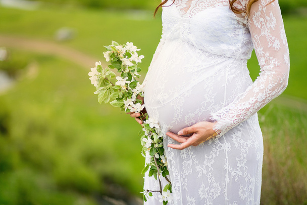Closeup of pregnant woman enjoying summer park, wearing long white dress, holding in hands bouquet of flowers, outdoors - Фото, изображение