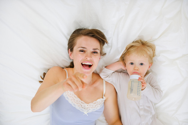 Mother laying with baby in bed and pointing in camera. Upper vie - Фото, изображение