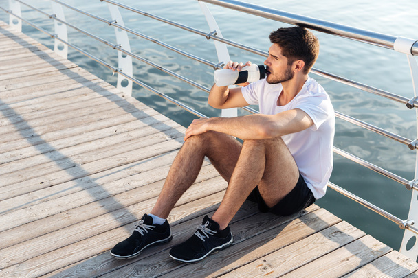 Male athlete sitting on pier and drinking water - Фото, зображення