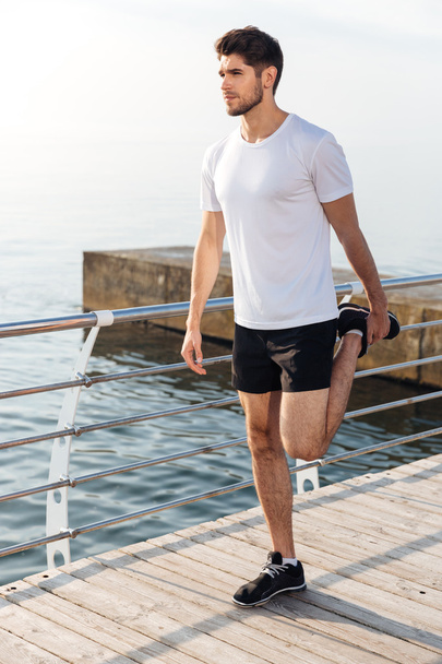 Sportsman doing exercises and stretching legs on pier near sea - Foto, afbeelding