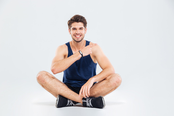 Smiling fitness man sitting with legs crossed and pointing away - Foto, afbeelding