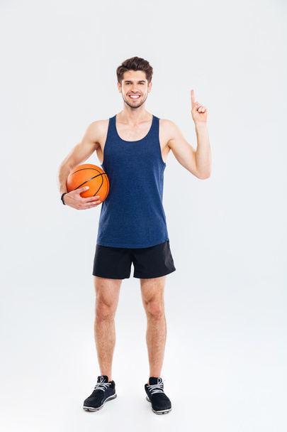 Smiling young sportsman holding basket ball and pointing finger up - Fotografie, Obrázek