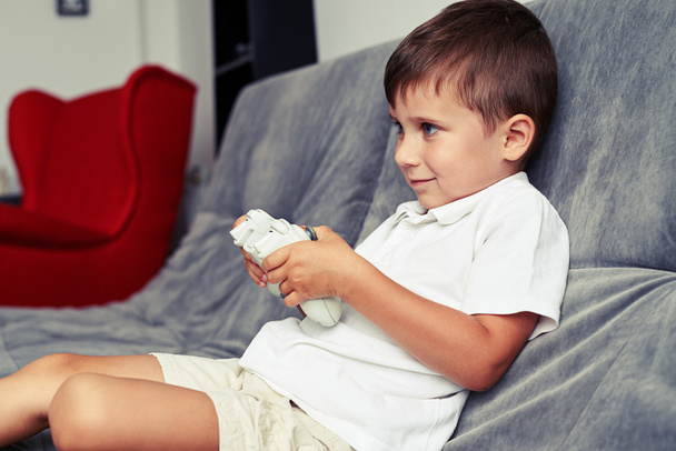 Small boy is rapt in playing a videogame sitting on grey sofa - 写真・画像