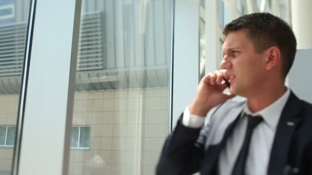 Handsome young businessman laughes while talking at phone and looking in modern building window - Materiał filmowy, wideo