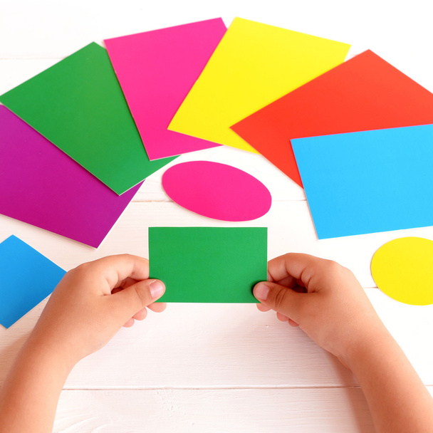 Το παιδί κατέχει πράσινο ορθογώνιο χαρτόνι στα χέρια και ψάχνει για το αντίστοιχο χρώμα κάρτα. Το παιδί μαθαίνει χρώματα και μορφή με χάρτινες κάρτες - Φωτογραφία, εικόνα