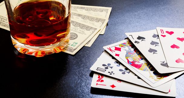 Glass of whiskey and playing cards and dollar bills on a black desk on the wooden table. - Valokuva, kuva