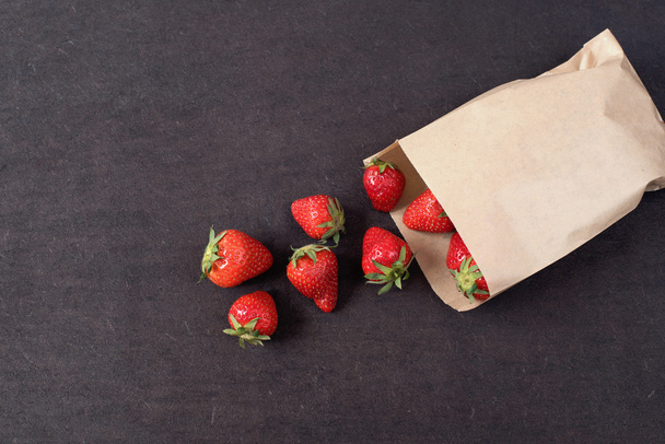 Paper bag with fresh red strawberries. Fresh strawberries in a small bag on a wooden style surface. Small group of strawberries on dark wood background with copy space - Photo, image