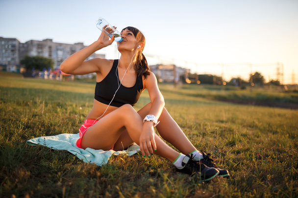 sportliche junge Frau trinkt Wasser. Mädchen sitzt auf Matte und hört Musik über Kopfhörer - Foto, Bild