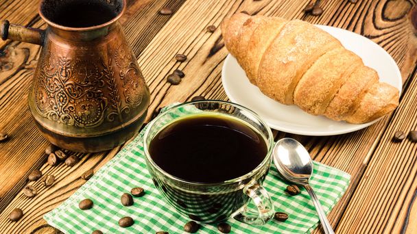 Чашка черного кофе на зеленой салфетке с круассаном, турецкий кофейник, деревянный стол в кафе
 - Фото, изображение