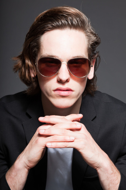Handsome young man with brown long hair and wearing white shirt and blue jacket and sunglasses isolated on grey background. Fashion studio shot. Expressive face. - Photo, Image