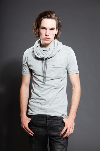 Handsome young man with brown long hair wearing grey shirt isolated on grey background. Fashion studio shot. Expressive face. - Photo, image