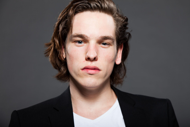 Handsome young man with brown long hair and wearing white shirt and blue jacket isolated on grey background. Fashion studio shot. Expressive face. - Foto, Imagen