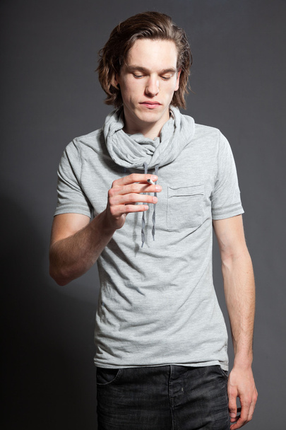 Handsome young man with brown long hair wearing grey shirt isolated on grey background. Smoking cigarette. Fashion studio shot. Expressive face. - Фото, изображение