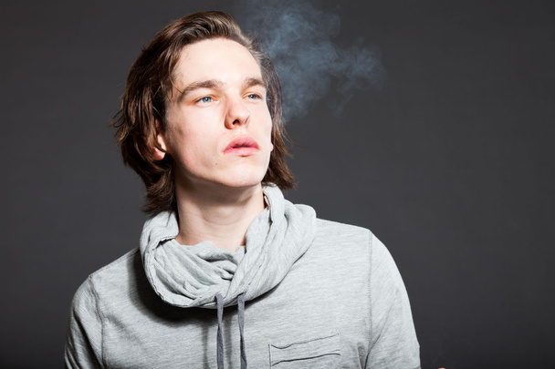 Handsome young man with brown long hair wearing grey shirt isolated on grey background. Smoking cigarette. Fashion studio shot. Expressive face. - Foto, afbeelding