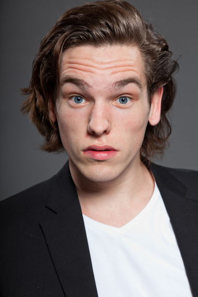 Handsome young man with brown long hair and wearing white shirt and blue jacket isolated on grey background. Fashion studio shot. Expressive face. - Фото, зображення