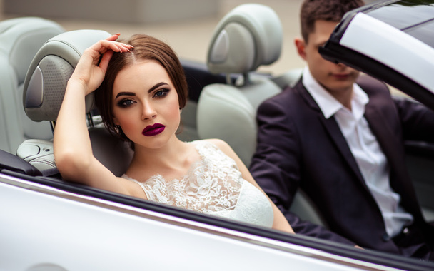 gorgeous bride with fashion makeup and hairstyle in a luxury wedding dress with handsome groom near white cabriolet car - Foto, imagen