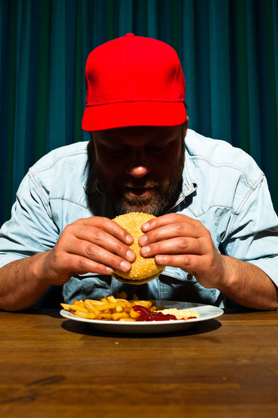 Hombre con barba comiendo comida rápida. Disfrutando de papas fritas y una hamburguesa. Camionero con gorra roja
. - Foto, imagen