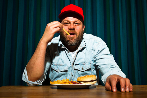 férfi szakállal eszik gyorséttermi étkezés. élvezi a sült krumpli és a hamburger. traktoros piros sapkát. - Fotó, kép