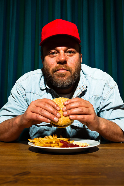 férfi szakállal eszik gyorséttermi étkezés. élvezi a sült krumpli és a hamburger. traktoros piros sapkát. - Fotó, kép