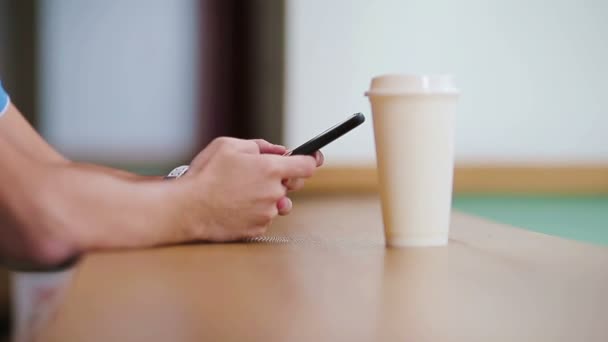 Primer plano de las manos masculinas sosteniendo el teléfono celular y el vaso de café en la cafetería. Hombre usando smartphone móvil. Un chico tocando una pantalla de su smarthone. Fondo borroso, horizontal
. - Imágenes, Vídeo
