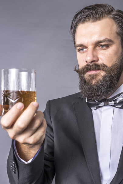 Egy szakállas, retró külsejű fiatalember portréja, aki egy pohár whiskyt tart a szürke háttér felett. - Fotó, kép