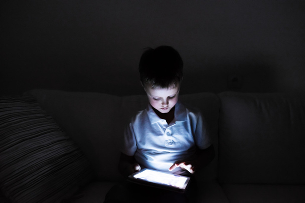 Мальчик, сидящий в темноте, играющий с планшетом - Фото, изображение